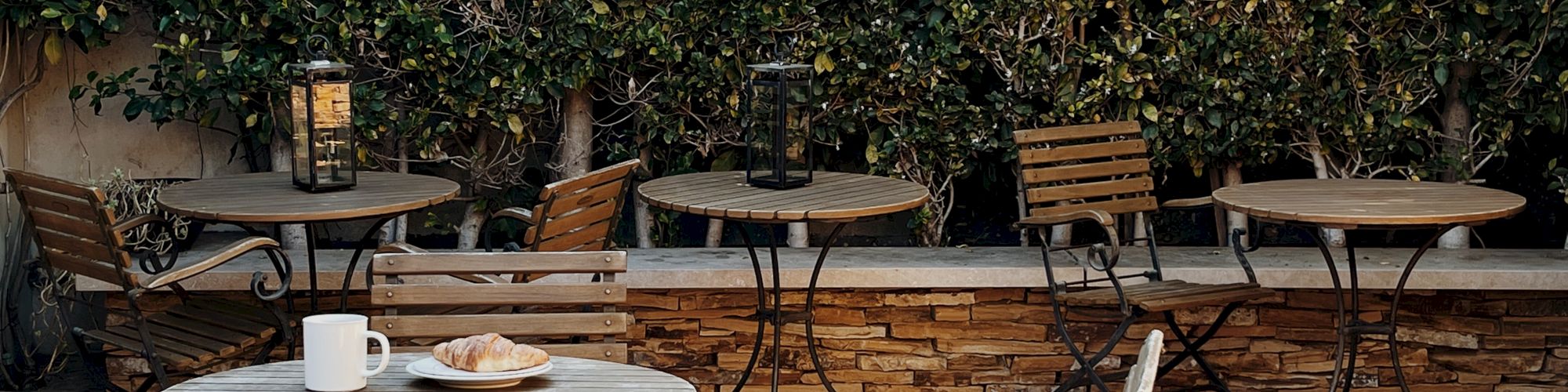 An outdoor seating area with metal tables and chairs is shown, surrounded by greenery in the background, and a coffee cup and pastry are on one table.